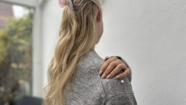 Schulterluxation: Schulter ausgekugelt | Radiologie Mühleninsel. Bild von junger Frau mit langen blonden Haaren, die sich an die rechte Schulter fasst von hinten. sie steht in einem wintergarten und hat eine graue strickjacke an Schulter ausgekugelt? Erfahren Sie, welche Symptome auf eine Schulterluxation hinweisen, und welche unsere Untersuchungen empfohlen sind.